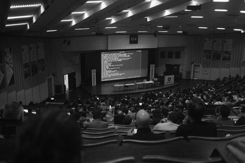 Black and white view of a full lecture hall, the audience in their seats and a speaker presenting