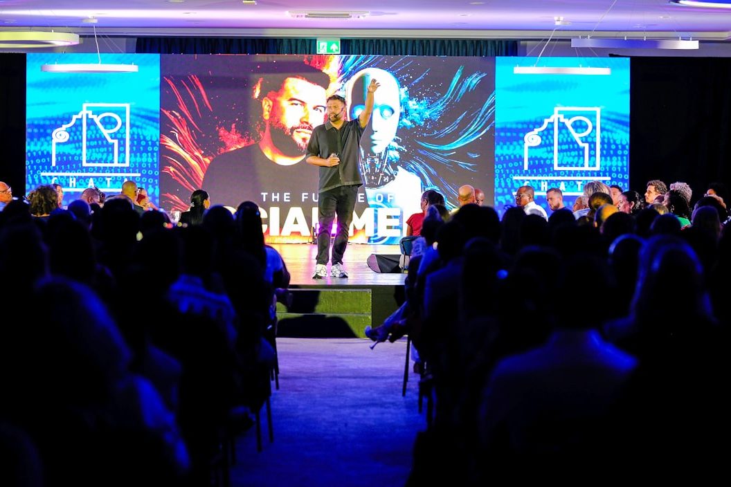 Speaker on stage before a large backdrop and branding screens with a seated audience at a tech conference